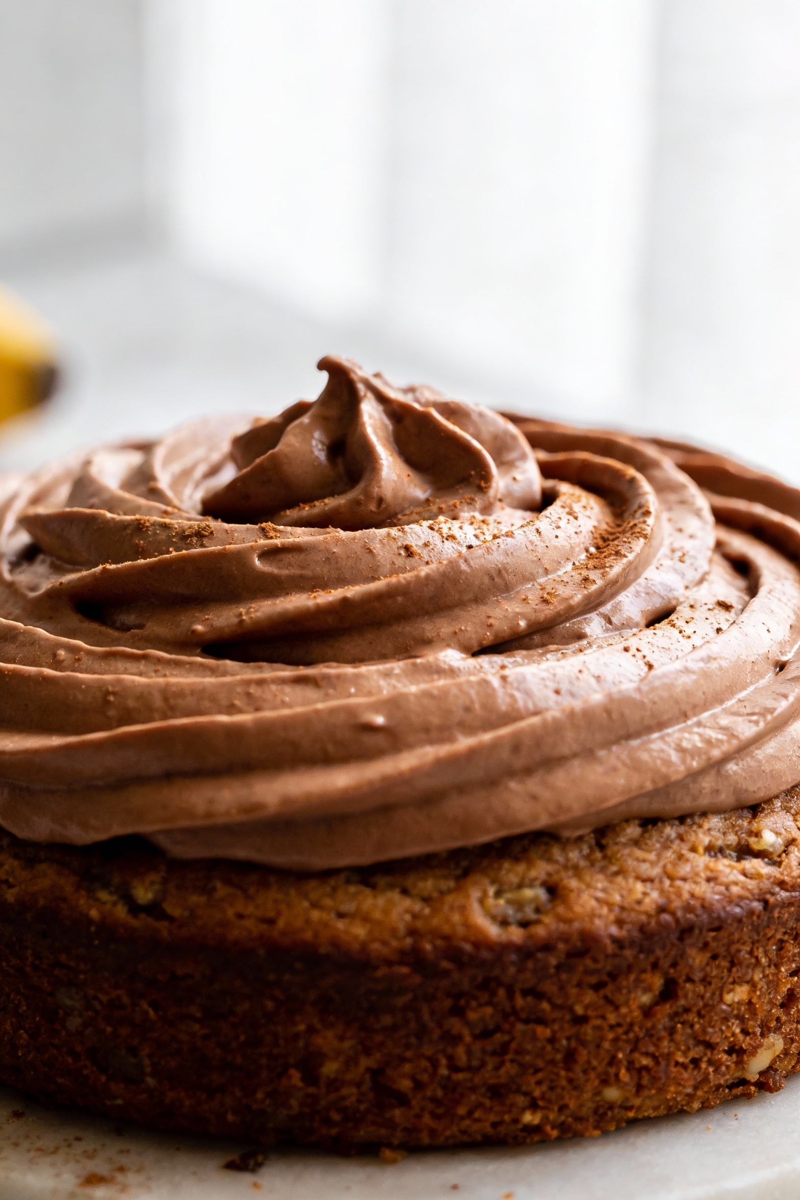 1. Close-up detail of a thick, glossy cocoa Greek-yogurt protein frosting swirled on a fully cooled healthy banana cake,