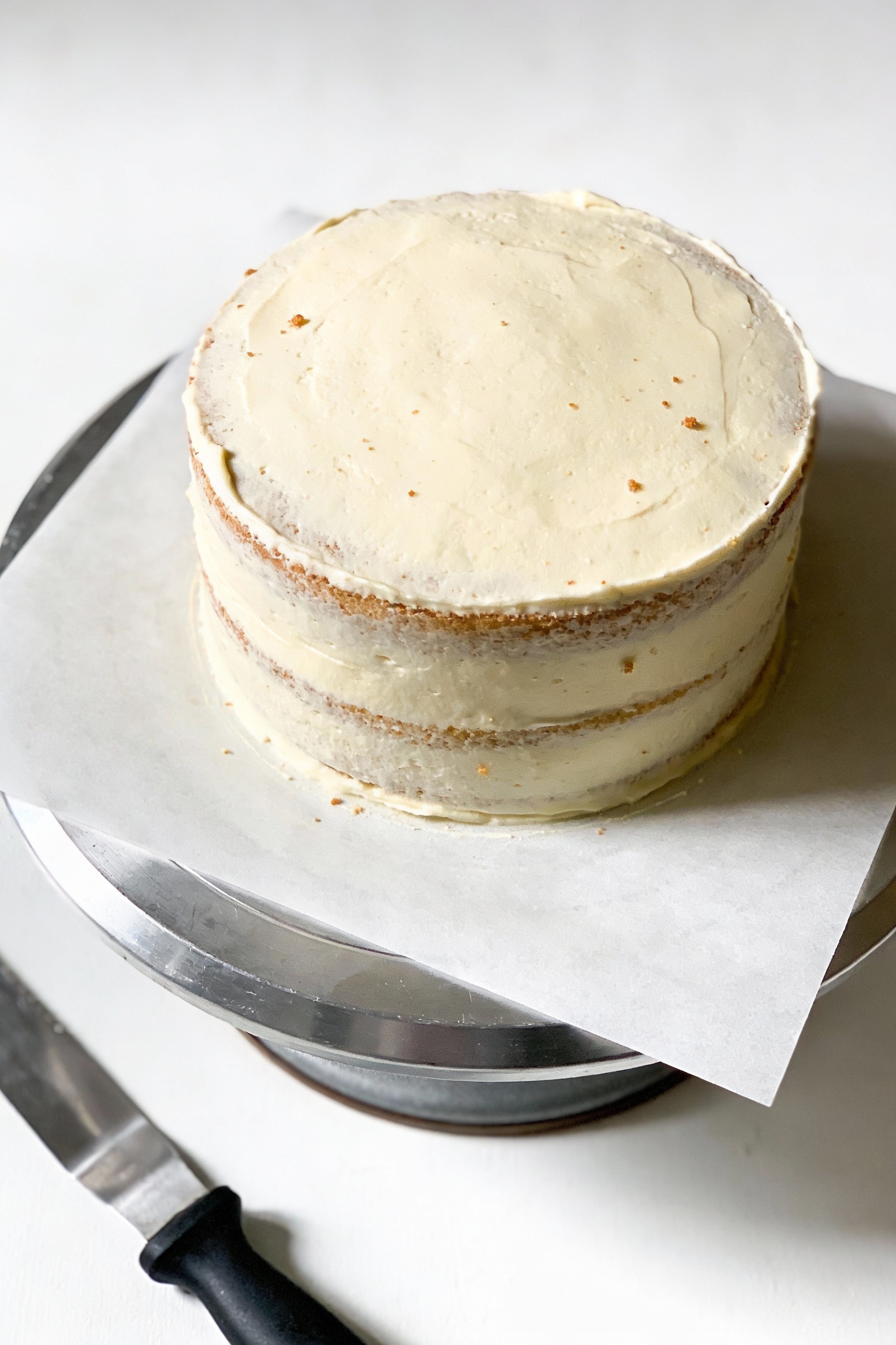 Overhead shot of a two-layer vanilla butter cake after crumb coat, thin pale frosting layer with a few visible crumbs an