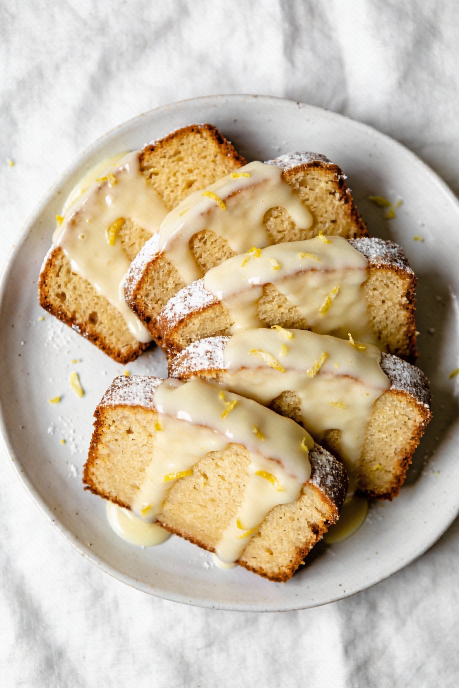 2. Overhead shot of lemon glaze loaf slices fanned on a white ceramic plate, shiny lemon glaze dripping over the cut edg