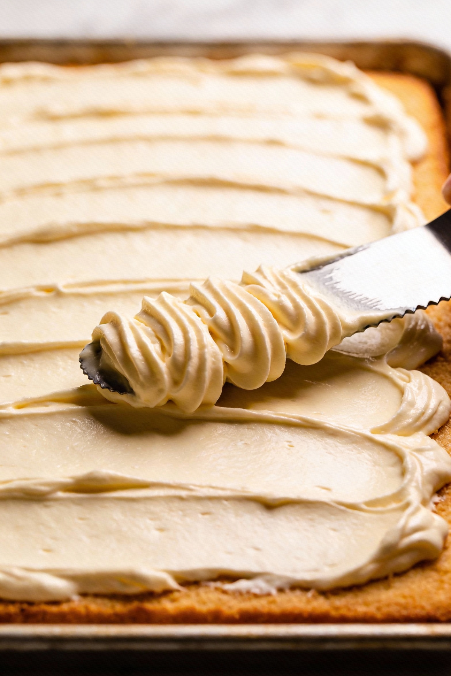Close-up detail of vanilla buttercream being spread in thick swoops over a fully cooled sheet cake with an offset spatul