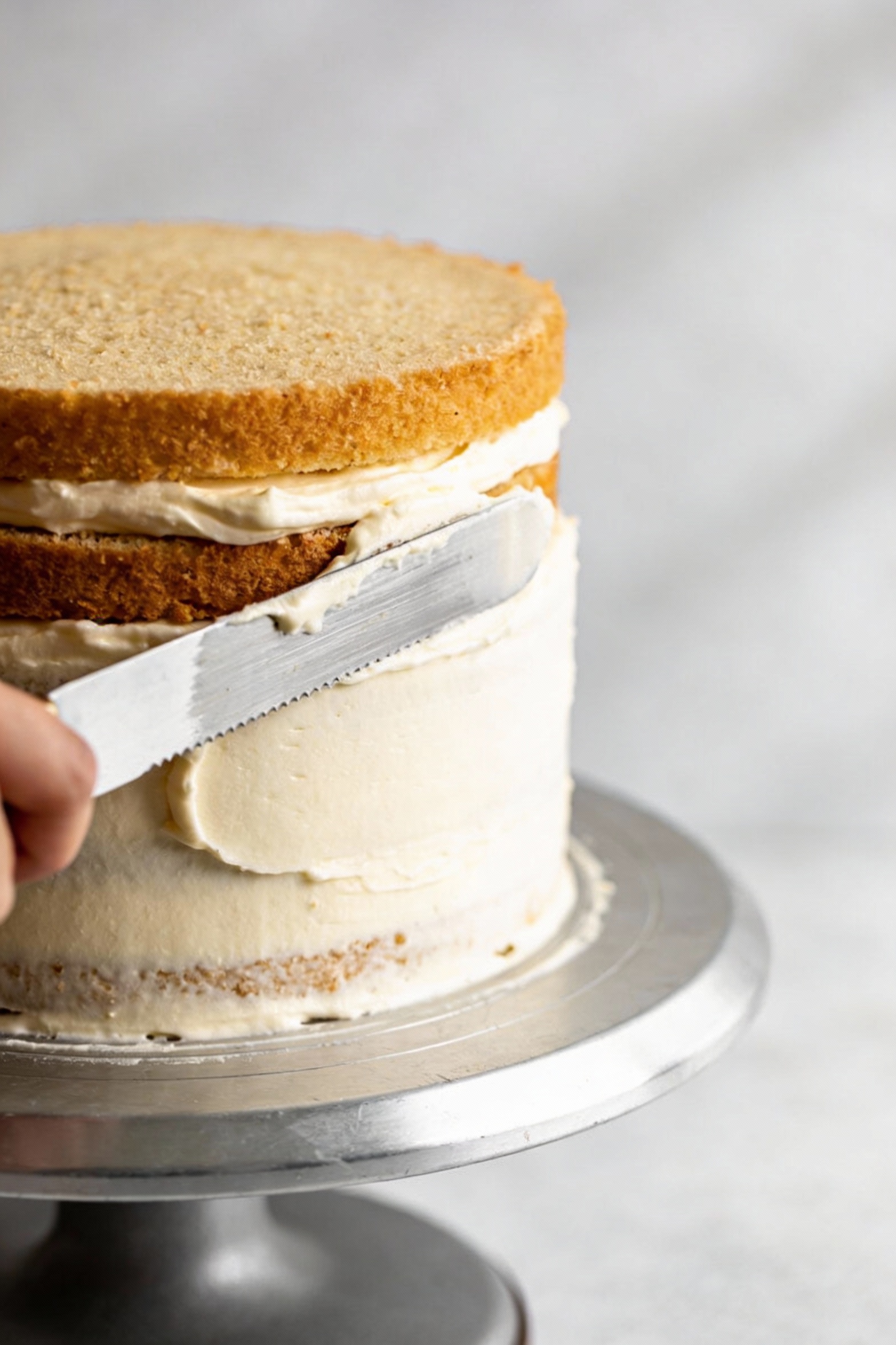 Vanilla celebration layer cake being assembled: two golden layers with a thin crumb coat of buttercream, offset spatula 