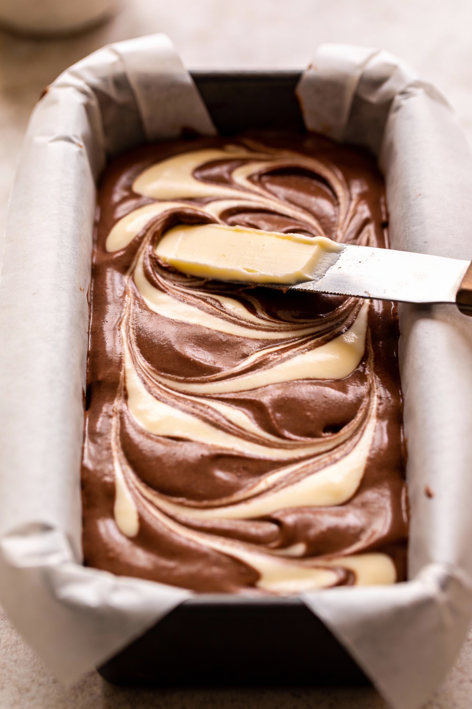 3. Cooking process: chocolate marble loaf batter in a 9x5 pan mid-prep, distinct vanilla and cocoa swirls being gently d