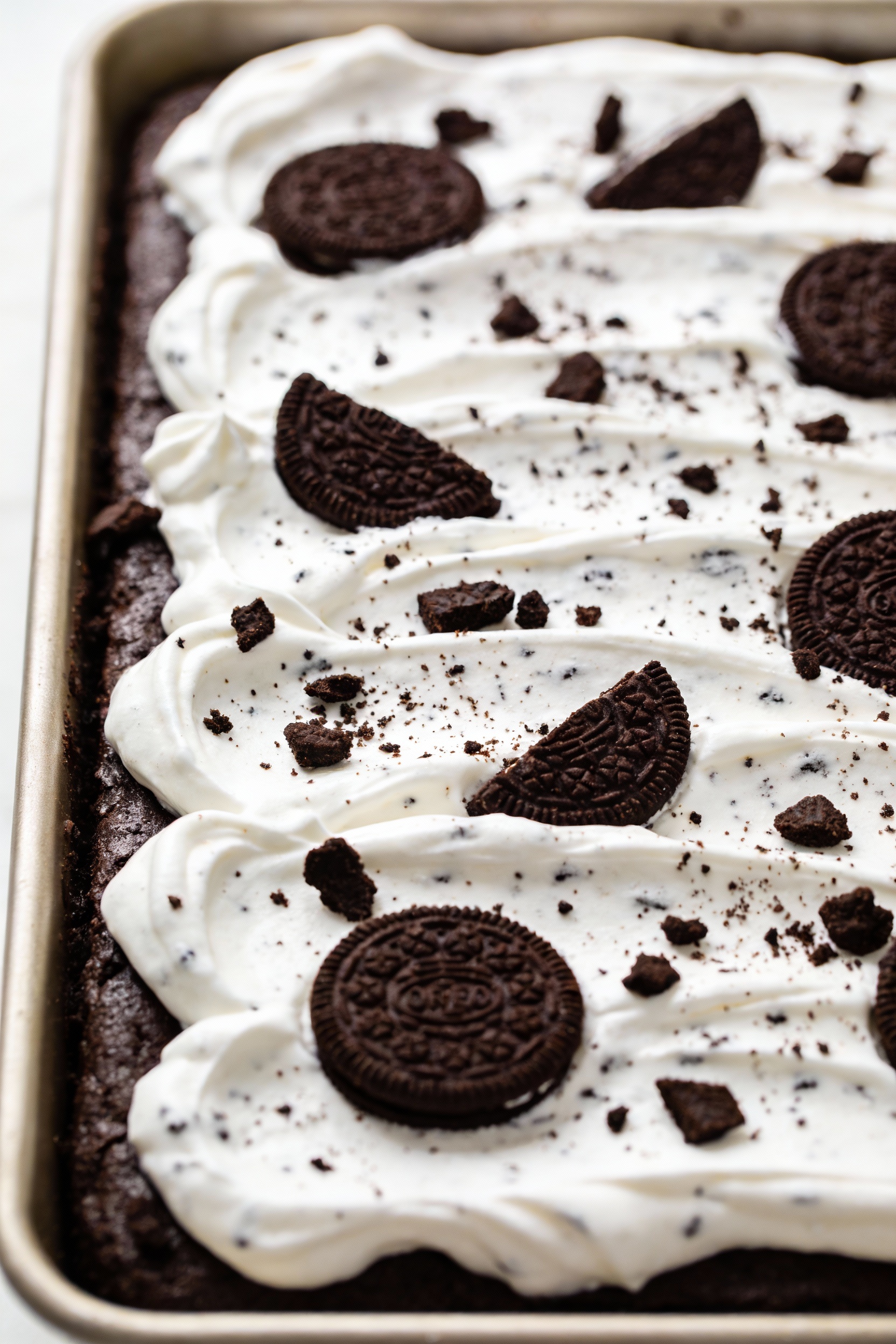 Cooking process: cookies-and-cream poke cake being finished in the pan with whipped topping being spread into smooth wav